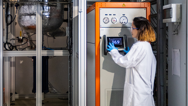 Das Bild zeigt eine Frau in Laborkleidung, die das Display einer verfahrenstechnischen Anlage bedient. 