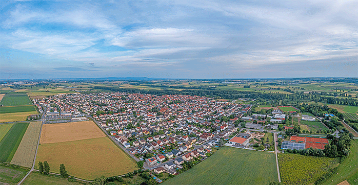 The picture shows a drone panorama over a village in the evening. Around the village are fields and forests. 