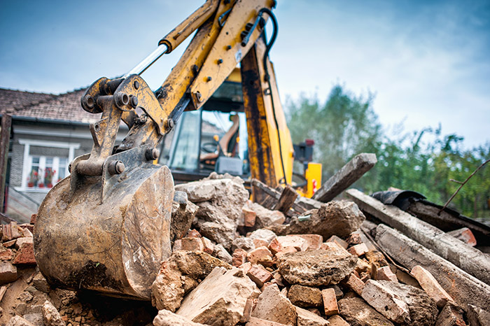The picture shows in the foreground an excavator whose shovel is lying on a pile of rubble. In the background you can see a house in the state of construction.