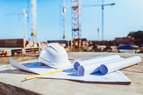The photo shows a close-up of a protective helmet lying on construction plans. You can also see a ruler. In the background you can see cranes and a construction site scenario.