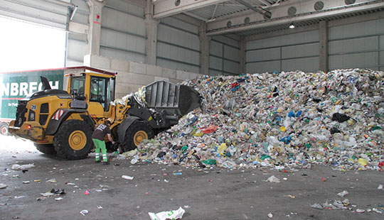 The picture shows an excavator driving plastic waste up to a pile of plastic garbage.