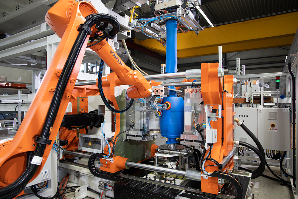 The photo shows orange robot arms reaching for a blue plastic barrel. The barrel is being cast from straight. An extruder can be seen at the top of the picture. At the moment the picture is taken, a blue plastic tube is being pressed out of the extruder and used in molding. 